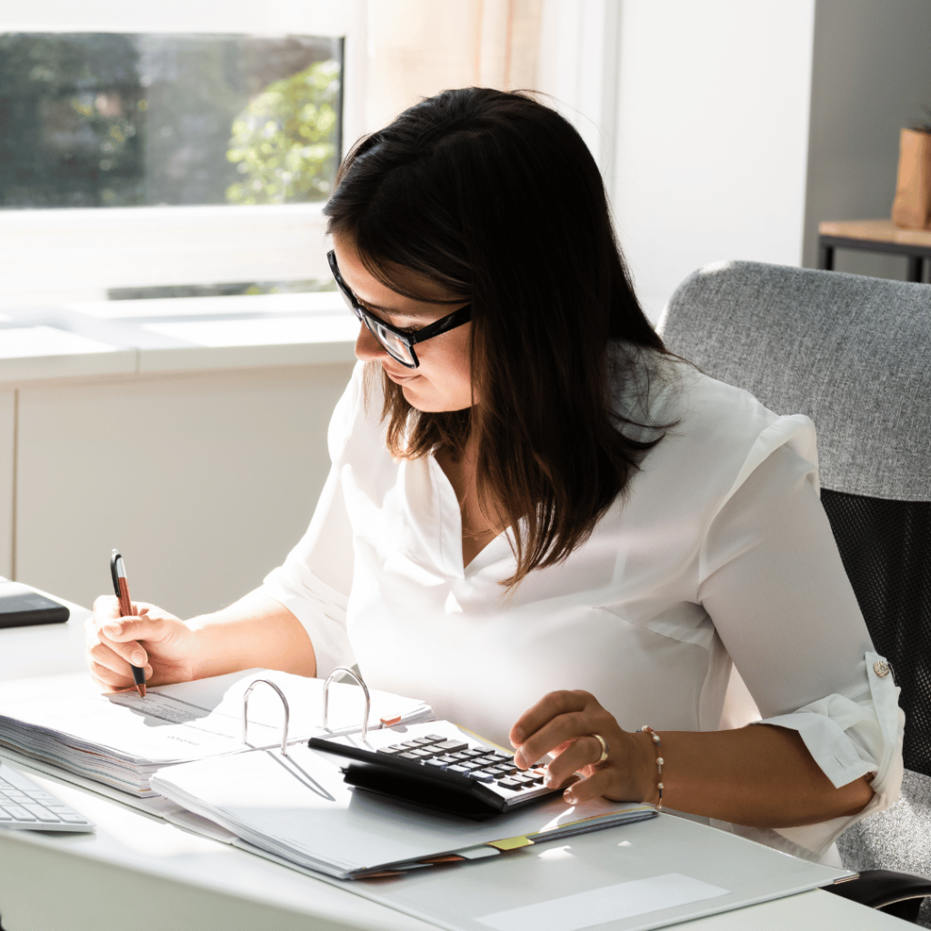 a lady accountant wearing white ling sleeve, sitting on her office desk with calculator in her hand and writing